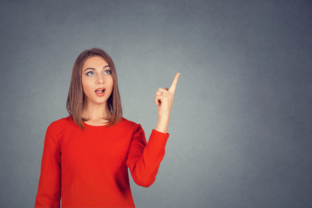 Surprised woman with open mouth pointing to the copyspace isolated on gray studio wall background. Attractive girl in casual clothes pointing her finger up. Business lady in red dress, bob hairstyle.の写真素材