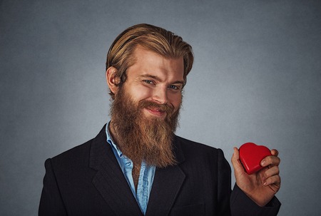 Young happy Caucasian bearded hipster businessman holding red heart shaped gift box ready for Valentine's day. Isolated on gray grey studio wall Background. Negative face expression, human emotion.の写真素材