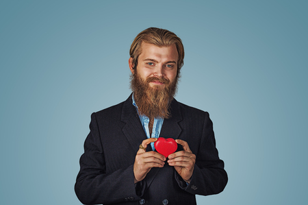 Handsome funny happy businessman in black jacket and blue shirt standing and holding red big heart shaped gift box looking at camera with good feelings. Indoor, studio shot isolated on blue backgroundの写真素材