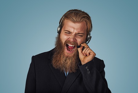 Angry Bearded hipster businessman aggressive customer support phone operator with headset, he is yelling and having a discussion with a customer isolated on blue Background. Negative face expression.の写真素材
