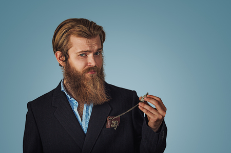 Bearded hipster Businessman looking skeptical holding retro pocket watch Isolated on blue Background. Negative face expression, human emotion, body language reaction attitude. Studio shot.の写真素材
