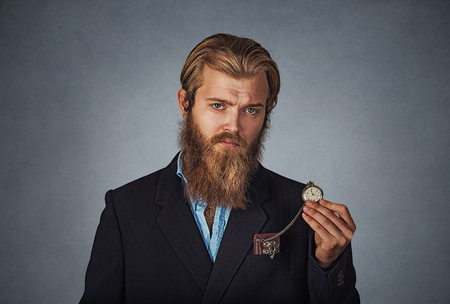 Time is ticking. Bearded hipster serious business man holding showing pocket watch Isolated on gray grey studio wall Background. Negative face expression, human emotion, body language, reaction.の写真素材