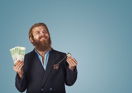Portrait of an adult Bearded hipster businessman cheerful positive man pumping fists standing isolated over blue studio wall background looking up holding money and pocket watch clock Isolated on blueの写真素材