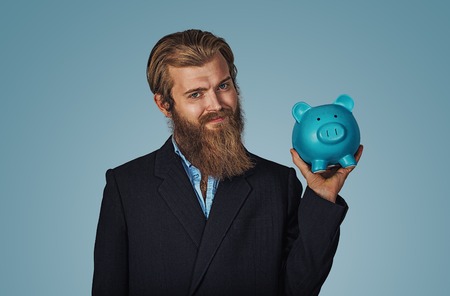Portrait of a smiling Bearded business man holding piggy bank money box Isolated on blue studio wall Background. Positive face expression human emotion body language reaction attitude. Horizontalの写真素材