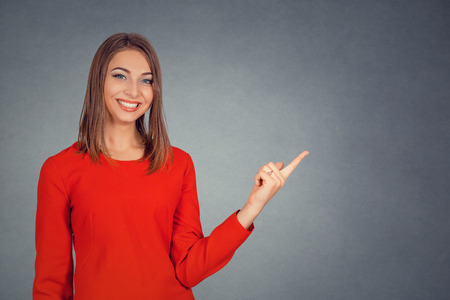 Smiling young woman showing pointing with finger at copy space for product or text. Business lady in red dress, bob hairstyle. Isolated on gray grey studio wall Background. Positive face expression.の写真素材