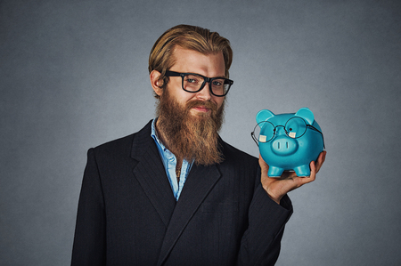 Closeup portrait head shot young serious smiling happy hipster businessman holding Piggy bank both wearing glasses isolated on gray studio wall background. Save money, savings on eyeglasses conceptの写真素材