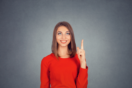 Look up here. Beautiful funny happy woman presenting showing copy space above head with index finger, aha eureka hand gesture. Business lady in red dress, bob hairstyle. Isolated on gray studio wallの写真素材