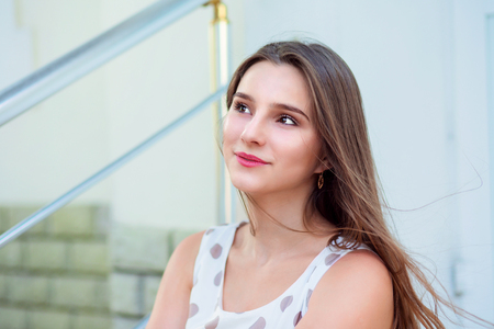 Teen girl with long hair sitting on steps looking up to side. Beautiful young girl in white dress with brown polka dots. Mixed race asian russian. Horizontal image outdoors home office on background.の写真素材