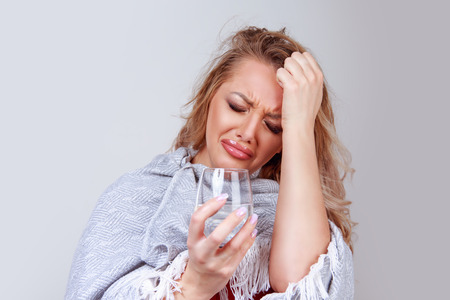 Pretty adult blonde woman feeling tired and hangover headache while holding glass of water on gray background in studio.の写真素材