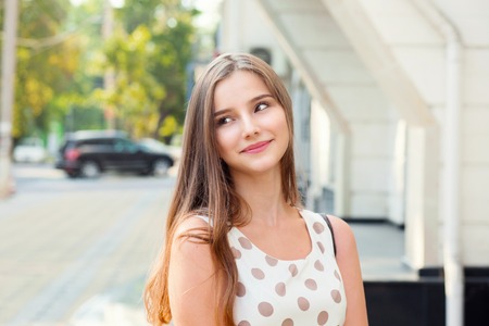 Teen girl with long hair smiling daydreaming looking up to side. Beautiful young girl in white dress with brown polka dots. Mixed race asian russian. Horizontal image outdoors home office background.の写真素材