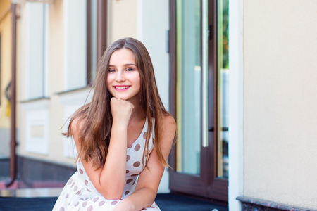Portrait of cheerful young woman sitting outdoors and smiling. Beautiful young girl in white dress with brown polka dots. Mixed race asian russian. Horizontal image outdoors home office on background.の写真素材