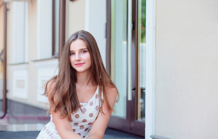 Teen girl with long brunette hair sitting on the marble steps. Beautiful young girl in white dress with brown polka dots. Mixed race asian russian. Horizontal image outdoors home office on background.の写真素材