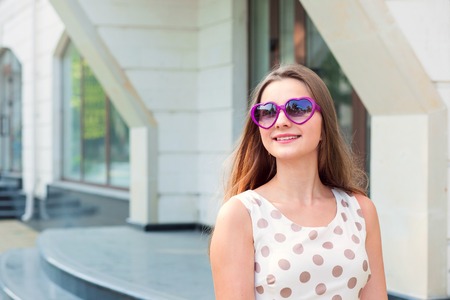 Teen girl with long brunette hair heart shaped purple sunglasses looking up to side to copy space dreaming outdoors home office on background. Beautiful young girl in white dress with brown polka dotsの写真素材