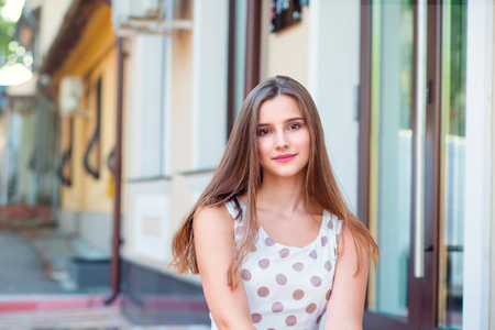 Teen girl with long brunette hair smiling sitting on steps. Beautiful young girl in white dress with brown polka dots. Mixed race asian russian. Horizontal image outdoors home office on background.の写真素材