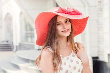 Beautiful young woman in hat walking in the city smiling looking to camera. Fashion girl. Beautiful young teenager in white dress with brown polka dots and pink hat on head. Mixed race asian russian.の写真素材