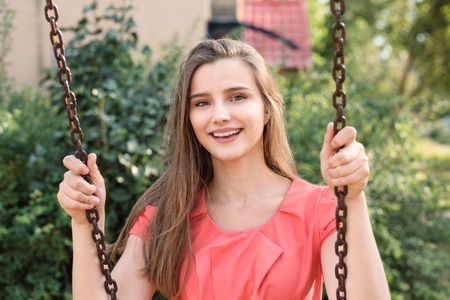Pretty happy brunette swinging smiling near house in park on a sunny day. Beautiful young girl in pink dress. Mixed race asian russian model. Horizontal image outdoors home, green bush on background.の写真素材