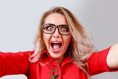 Excited blonde woman in glasses with wide-opened eyes and mouth opened taking selfie and looking at camera on gray background.の写真素材