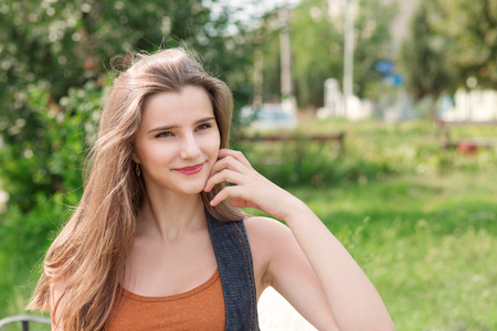 portrait of a satisfied woman looking away to side leaning on a bench with an rustic green background. Beautiful young teen in orange shirt jeans jacket.  Mixed race asian russian girl Horizontal shotの写真素材
