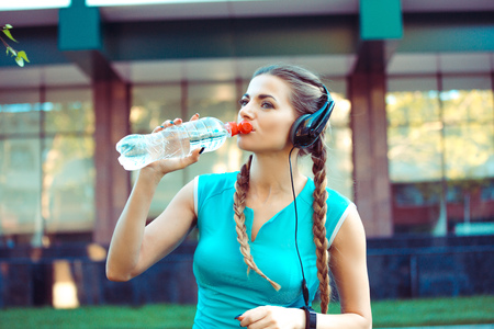 Modern young sportswoman wearing headphones and drinking water from bottle on streetの写真素材