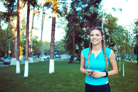 Excited young sportive woman with braids holding phone and smiling away in green parkの写真素材