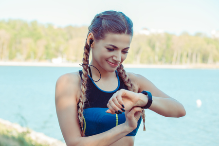 Modern slim sportive woman in headphones using smart watch while standing outdoorsの写真素材
