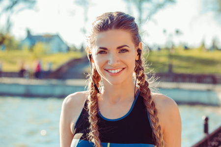 Wonderful young fit sportswoman with braids wearing headphones and smiling at camera in sunlightの写真素材