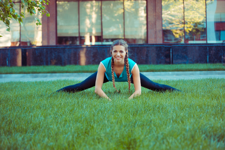 Pretty young brunette in sportswear stretching legs on green meadow standing in splits and smiling at cameraの写真素材