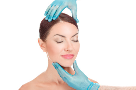 Beautiful Woman after Plastic Surgery Operation Cosmetology. Doctor hands around patient face. Portrait of attractive caucasian woman brunette isolated on white background, horizontal studio shot.の写真素材