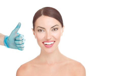 Young brunette woman smiling, looking at you camera happy, doctor hand in blue glove showing thumb up like hand gesture, that she is looking great isolated on white background. Beautify concept.の写真素材