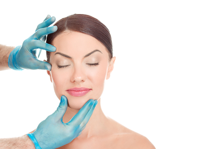 Woman with eyes closed after aesthetic surgery, smiles while the doctor shows the results isolated on white background with copy space. Doctor hands around patient face. Beautify conceptの写真素材
