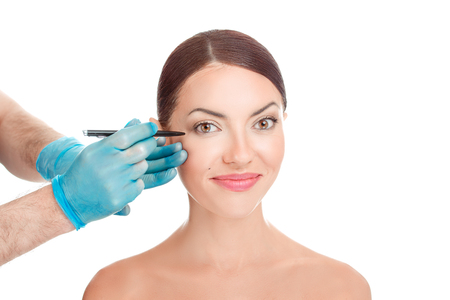 Plastic surgeon about to draw surgery line under eye of girl. Hand in blue glove holding pencil. Plastic surgery, beauty portrait, closeup  isolated on white background with copy space. Beautifyの写真素材