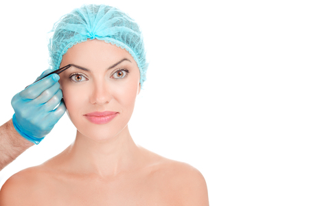 Young woman having eyebrows hair plucked by doctor hand closeup portrait isolated on a white background. Beauty studio shot. Beautician plucking brows depilating with tweezers. Trimming brow in salon.の写真素材