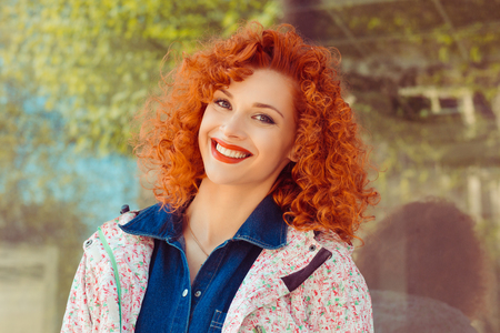Happy cheerful young woman with curly redhead ginger hair rejoicing at positive news, looking at camera with joyful and charming smile. Ginger student girl relaxing indoors after collegeの写真素材