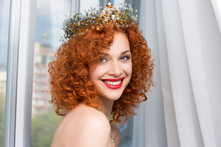 Happy bride. Beautiful red head curly woman with crown fashion model person girl looking at you camera smiling at boyfriend groom isolated  near window indoor white curtains background. Miss sunshineの写真素材