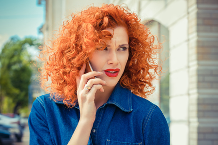 What are you saying. Portrait angry frustrated, red curly hair woman talking on mobile phone standing outside with city on background. Negative emotions face expression reaction feelings.の写真素材