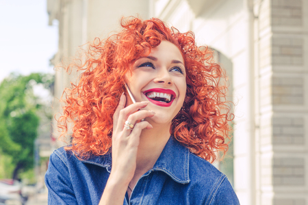 Beautiful red curly hair woman talking on mobile phone, laughing, looking upwards. Isolated in the city. Positive human emotion facial expression feeling.の写真素材