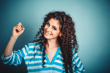 Portrait closeup of happy beautiful young woman writing holding pen looking at you camera isolated blue wall background with copy space. Human face expression emotion feeling body languageの写真素材