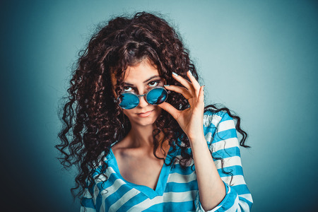 Headshot serious angry bitchy curly woman wife holding sunglasses down skeptically looking at you isolated blue wall background, blue shirt. Human face expression body language, attitude, perceptionの写真素材