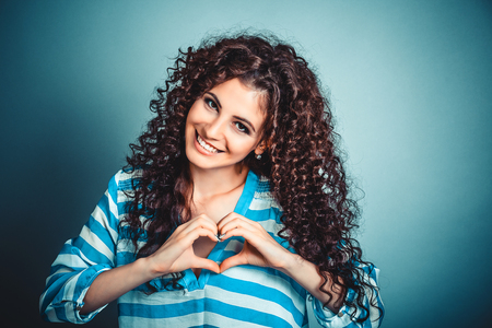 Closeup portrait smiling cheerful happy young curly girl woman making heart sign with hands isolated on blue wall background. Positive human emotion expression feeling life perception body languageの写真素材