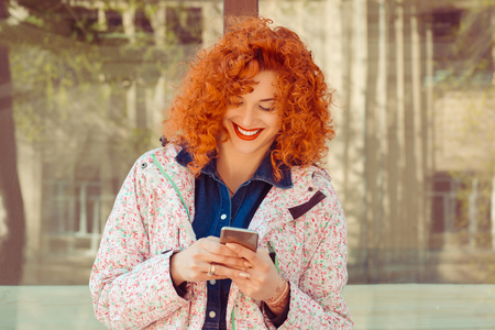 Close up portrait of beautiful happy young redhead american girl woman reading text message on mobile phone in a bus station outdoors smiling. Positive human emotions face expression feeling.の写真素材