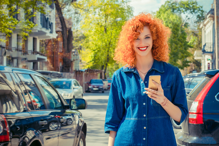 Taxi app. Pretty happy smiling urban woman using smart phone, standing on city street on sunny day waiting for taxi. Beautiful redhead girl holding mobile, texting sms message wearing jeans shirtの写真素材