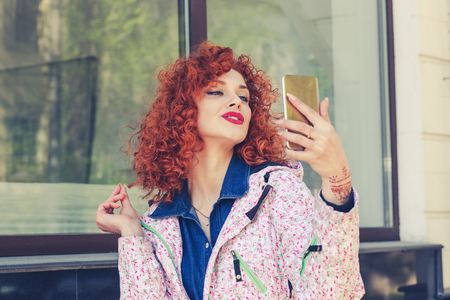 Portrait of stylish young redhead curly woman near her apartment complex outdoors taking selfie with her smart phoneの写真素材