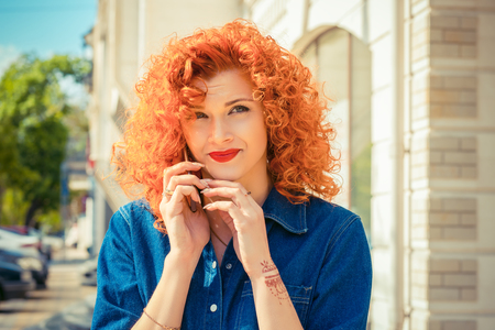 Annoyed woman with red curly hair talking to mobile phone, bored by the conversation looking up to side frustrated standing outside isolated city office building background. Negative emotionの写真素材