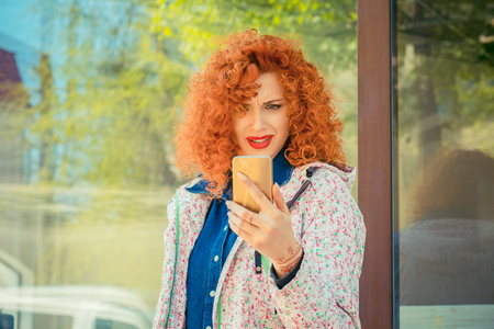 Portrait of stressed office young frustrated woman holding cellphone in hand on the city street in summer, looking at screen with cross face expression, mad at stressful texts and callsの写真素材