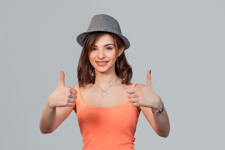 Happy smiling woman with gray hat showing thumbs up like gesture isolated grey studio wall background. Positive emotion face expression sign symbol body languageの写真素材