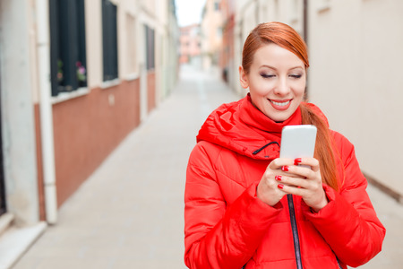 Tourist using internet. Front view portrait of a happy lady using a smart phone in the street, Murano Island, Venice Italy. Model in red winter coat clothing redhead hair standing on street backgroundの写真素材