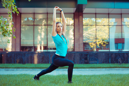 Pretty young brunette in sportswear stretching legs and hands on green meadow standing in splits looking away on a city urban backgroundの写真素材