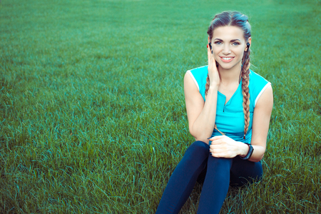 Happy young athletic woman wearing headphones listening to music sitting on a green grass lawn outdoors, outside in a summer parkの写真素材