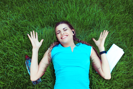 From above portrait of sporty euphoric woman hands in air happy gesture lying down a green grass meadow lawn outside in the city. Girl  with bottle of water, towel around, wins in competition sport.の写真素材