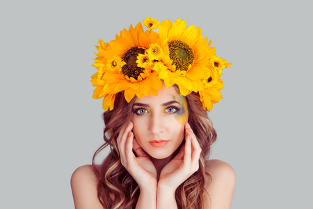Beautiful young woman wearing floral headband isolated on gray background. Yellow sunflowers wreath of flowers on head looking at you smiling slightly hands touching face, posing cute gorgeous girl.の写真素材